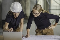 <p>Front view of two women standing by the desk full of blueprints and laptop, 